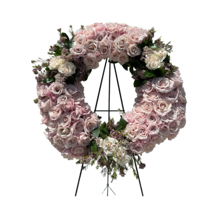 A sophisticated sympathy wreath displayed on a black metal easel, featuring a full ring of blush and ivory roses accented with light pink waxflower and soft greenery. This circular floral tribute conveys peace, remembrance, and eternal love—ideal for funeral services, memorials, or celebrations of life with a refined and gentle aesthetic.