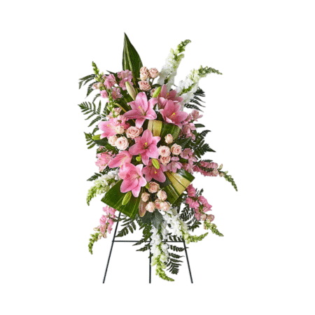 An elegant standing funeral spray displayed on a black easel, composed of pink lilies, soft blush spray roses, white snapdragons, and cascading ferns. Large tropical greens and folded ti leaves add structural contrast and depth. This tall and graceful floral tribute conveys heartfelt sympathy, support, and love—ideal for memorials, funerals, or celebrations of life.