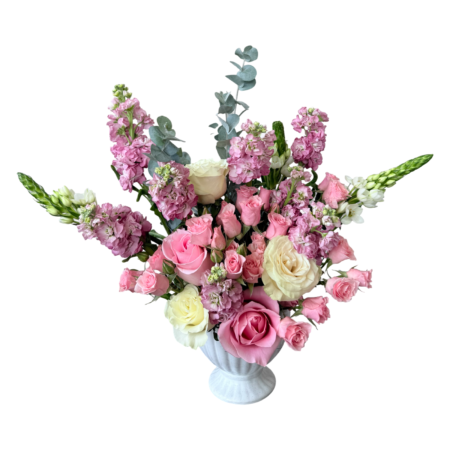A soft and elegant floral arrangement displayed in a white ceramic fluted urn-style vase. The bouquet features a romantic blend of pale pink roses, ivory roses, blush spray roses, lavender stock, white snapdragons, and eucalyptus stems. The airy, garden-inspired design and pastel tones make it ideal for both classic celebrations and gentle sentiments.