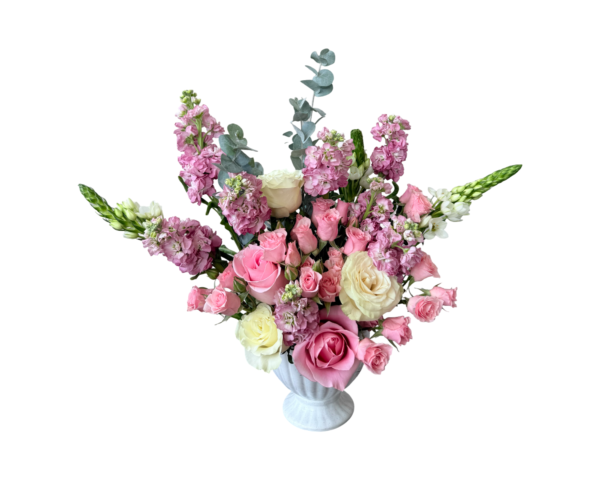 A soft and elegant floral arrangement displayed in a white ceramic fluted urn-style vase. The bouquet features a romantic blend of pale pink roses, ivory roses, blush spray roses, lavender stock, white snapdragons, and eucalyptus stems. The airy, garden-inspired design and pastel tones make it ideal for both classic celebrations and gentle sentiments.