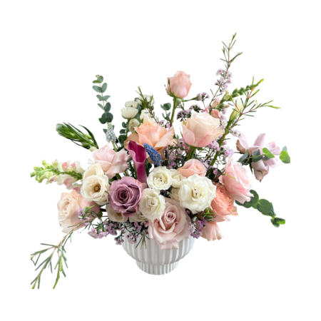 An ethereal garden-inspired floral arrangement designed in a fluted white ceramic vase, featuring soft blush roses, ivory spray roses, lavender roses, pale peach blooms, white ranunculus, pink waxflower, blue muscari, burgundy calla lilies, and touches of eucalyptus. With its natural flow and whimsical textures, this bouquet evokes a fresh spring meadow—perfect for refined gifting and serene occasions.