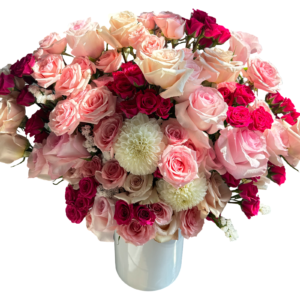 A lush and voluminous floral arrangement overflowing with shades of blush, pale peach, soft pink, and hot pink roses, accented by creamy white dahlias and set in a glossy white cylinder vase. The rich mix of full-sized roses and petite spray roses creates a classic, romantic silhouette that’s ideal for grand gestures, luxury gifts, and special milestones.