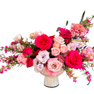 A vibrant floral centerpiece in a rounded marble-like pedestal vase, featuring a striking palette of hot pink, blush, and lavender tones. The arrangement includes bold red roses, soft pink roses, lavender roses, pink waxflower, peach spray roses, mauve chrysanthemums, pink stock, gerbera daisies, and textural filler greenery. A modern-meets-romantic design ideal for celebrations of love, power, and elegance.