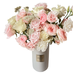 A soft and romantic floral arrangement presented in a tall white ceramic vase with a gold L’amour label. The bouquet features blush pink roses, ivory roses, white spray roses, baby’s breath, and white carnations. This airy and pastel-toned composition brings timeless elegance and tenderness, perfect for heartfelt gifting or meaningful celebrations.
