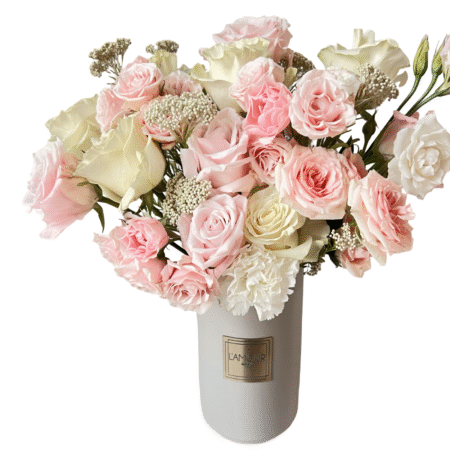 A soft and romantic floral arrangement presented in a tall white ceramic vase with a gold L’amour label. The bouquet features blush pink roses, ivory roses, white spray roses, baby’s breath, and white carnations. This airy and pastel-toned composition brings timeless elegance and tenderness, perfect for heartfelt gifting or meaningful celebrations.