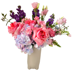 This elegant and feminine floral arrangement is designed in a tall, sculpted beige vase and features a romantic mix of soft pinks, lavenders, and purples. The bouquet includes large garden-style pink roses, pale pink lisianthus, lavender snapdragons, light purple hydrangeas, pink spray roses, and bold purple stock. The blend of textures and gentle tones gives the arrangement a dreamy, graceful feel, perfect for thoughtful occasions, romantic gestures, or celebratory events.