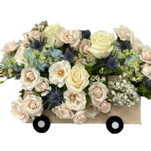 This whimsical and elegant floral design is arranged inside a wooden truck-shaped container, giving it a playful yet heartfelt touch. The bouquet features creamy ivory roses, blush spray roses, white ranunculus, pale blue hydrangeas, blue eryngium (thistle), wax flower, white baby’s breath, and soft green moss for texture. The gentle pastel tones combined with the unique presentation make this a charming choice for new beginnings, gentle condolences, or meaningful celebrations.