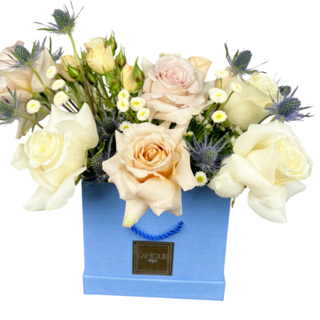 A stylish floral arrangement displayed in a powder-blue square gift bag from L’amour Designs. This elegant bouquet features cream and champagne roses, light peach spray roses, chamomile daisies, and blue eryngium thistle, blending soft neutrals with subtle pops of blue for a contemporary and gender-neutral gift. Ideal for elegant celebrations, corporate gifting, or welcoming a new baby boy.