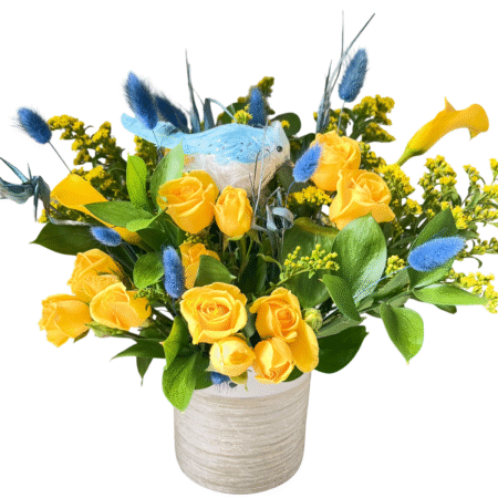 A bright and playful floral arrangement in a white textured ceramic vase featuring vivid yellow roses, yellow calla lilies, golden solidago (yellow filler), and blue dried bunny tails. Accented with a decorative artificial blue bird nestled among the flowers, this design bursts with contrasting sunny and sky tones—perfect for joyful celebrations, spring gifting, or gender reveals.