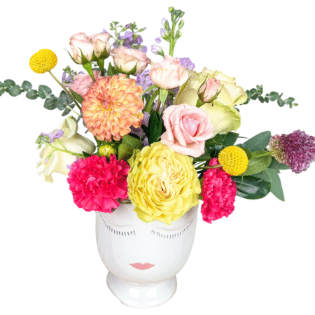 A whimsical and colorful floral arrangement displayed in a white ceramic vase with a serene face design. This bouquet includes soft pink roses, pale yellow garden roses, hot pink carnations, peach dahlias, yellow billy balls (craspedia), lavender stock, eucalyptus stems, and purple allium, accented with seasonal greenery. The design features a playful, artistic aesthetic with a balanced mix of bold and pastel tones—perfect for modern gifting and special occasions.