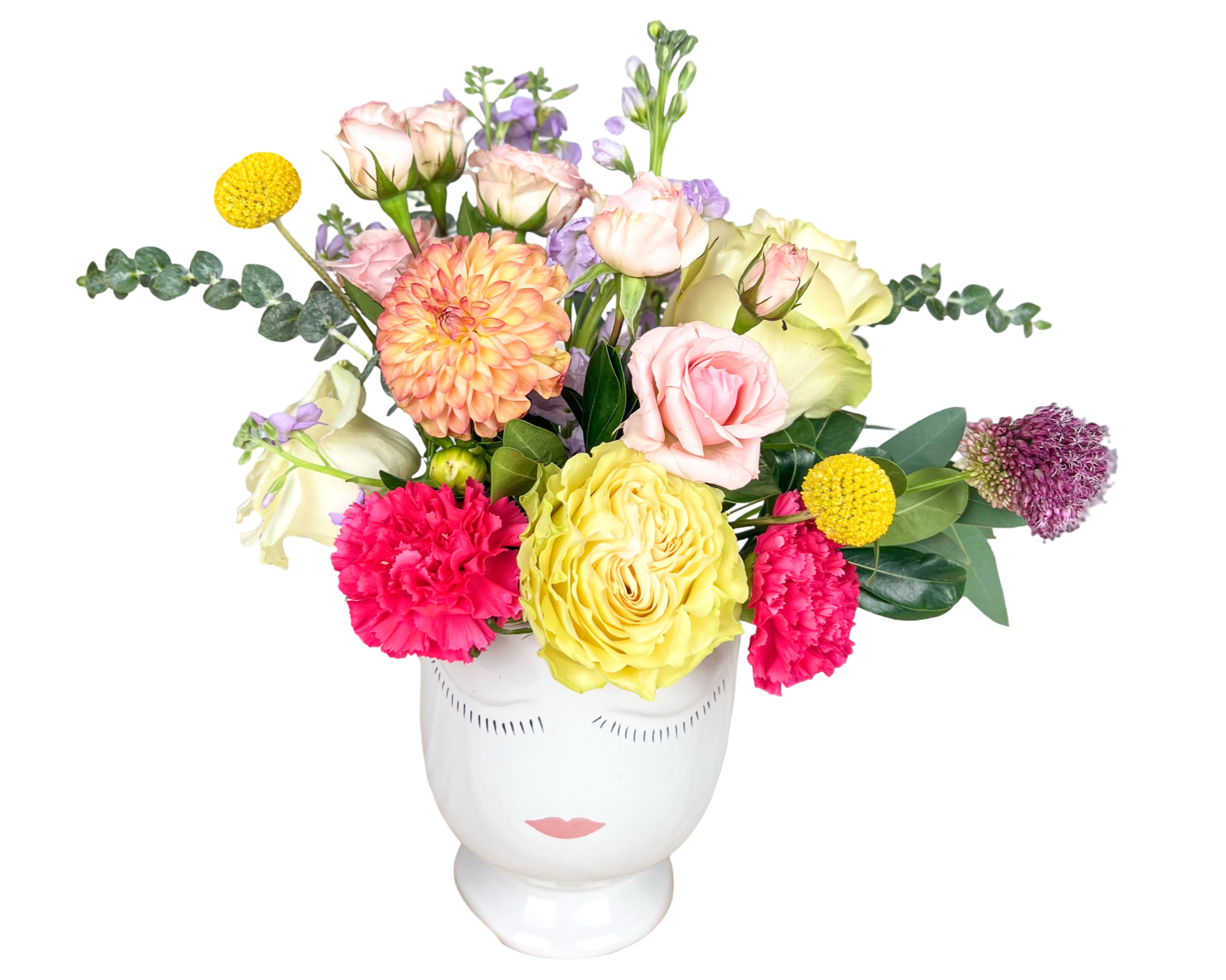 A whimsical and colorful floral arrangement displayed in a white ceramic vase with a serene face design. This bouquet includes soft pink roses, pale yellow garden roses, hot pink carnations, peach dahlias, yellow billy balls (craspedia), lavender stock, eucalyptus stems, and purple allium, accented with seasonal greenery. The design features a playful, artistic aesthetic with a balanced mix of bold and pastel tones—perfect for modern gifting and special occasions.