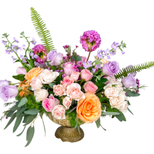 A garden-inspired floral arrangement in a vintage gold pedestal vase, bursting with lavender roses, peach and blush spray roses, creamy white roses, pink waxflower, deep magenta carnations, purple stock, and lush greenery including eucalyptus and ferns. This airy and artistic bouquet exudes a graceful charm and romantic feel, ideal for spring celebrations and elevated gifting.