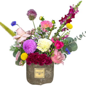 A vibrant and eclectic floral arrangement presented in a rustic stone-textured L’amour Designs container. This design features a striking mix of textures and colors, including white roses, fuchsia spray roses, lavender roses, yellow craspedia (billy balls), purple dahlias, burgundy coxcomb, blue cornflowers, pink lisianthus, burgundy snapdragons, pink dahlias, allium, and various greenery including eucalyptus and grasses. A playful, garden-style piece perfect for creative gifting or joyful occasions.