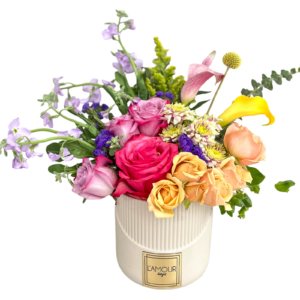 A joyful mixed floral arrangement in a ribbed ivory ceramic vase from L’amour Designs, featuring a lively palette of yellow, pink, purple, lavender, and peach tones. The bouquet includes hot pink roses, lavender roses, peach roses, yellow mini roses, yellow calla lilies, pink calla lily, purple statice, solidago, craspedia (billy ball), variegated mums, and lavender stock, arranged with a modern garden flair.